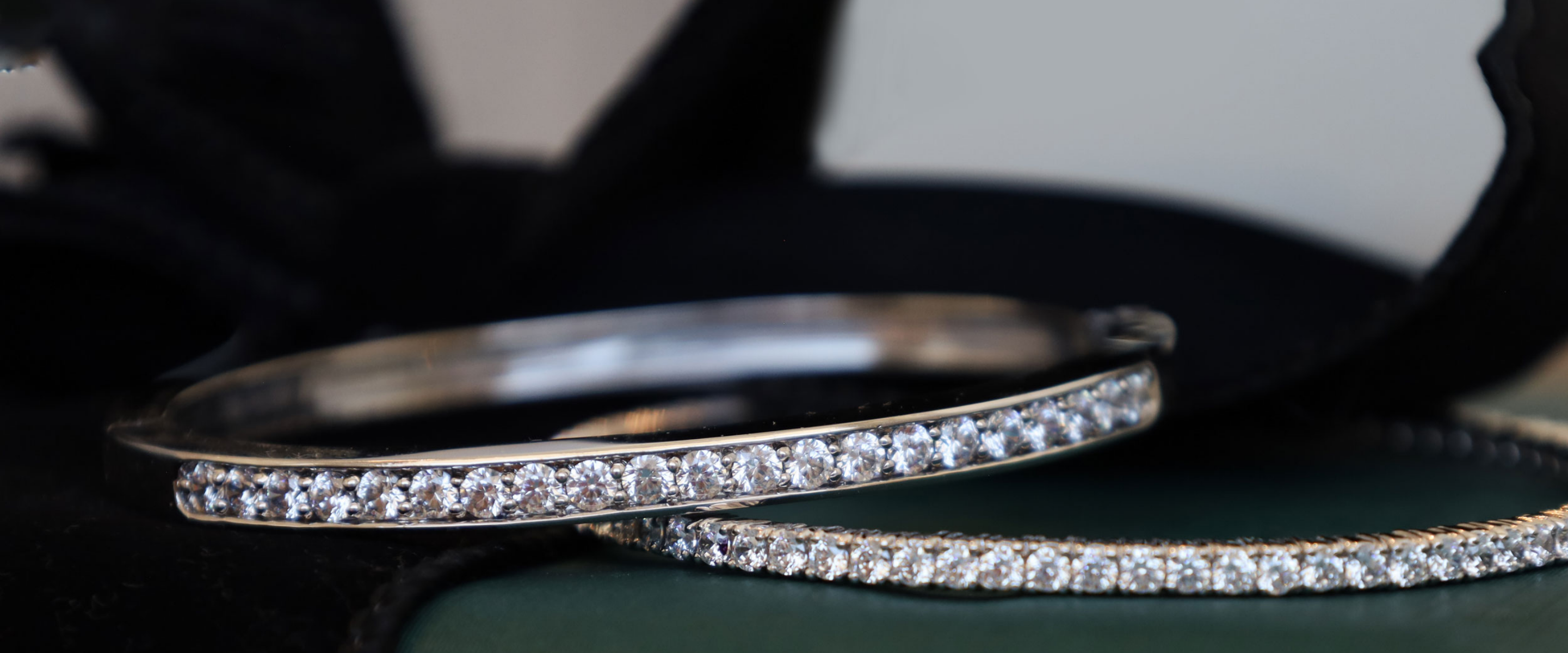 Two slim silver-toned bangle bracelets, each set with a row of clear stones, resting on a dark green surface with a black velvet box in the background.