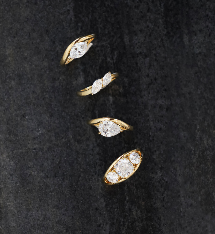 Four yellow gold rings with marquise-cut and oval-cut diamonds arranged diagonally on a dark textured background.