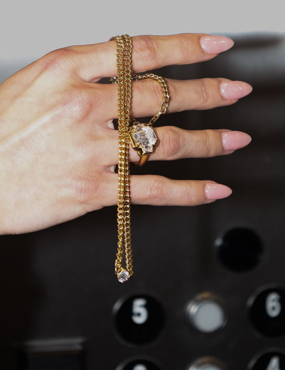 A gold chain necklace with a large clear gemstone pendant draped over a hand, with a matching gold ring on the middle finger, set against a dark elevator-style panel background.
