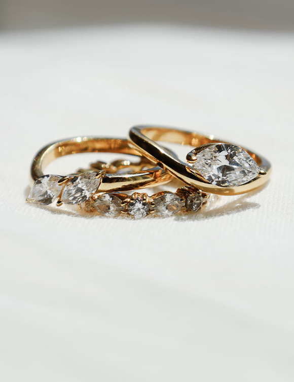 A close-up of three gold rings on a white textured surface. One ring features a large oval clear gemstone, while the other two are adorned with smaller marquise-cut clear gemstones.