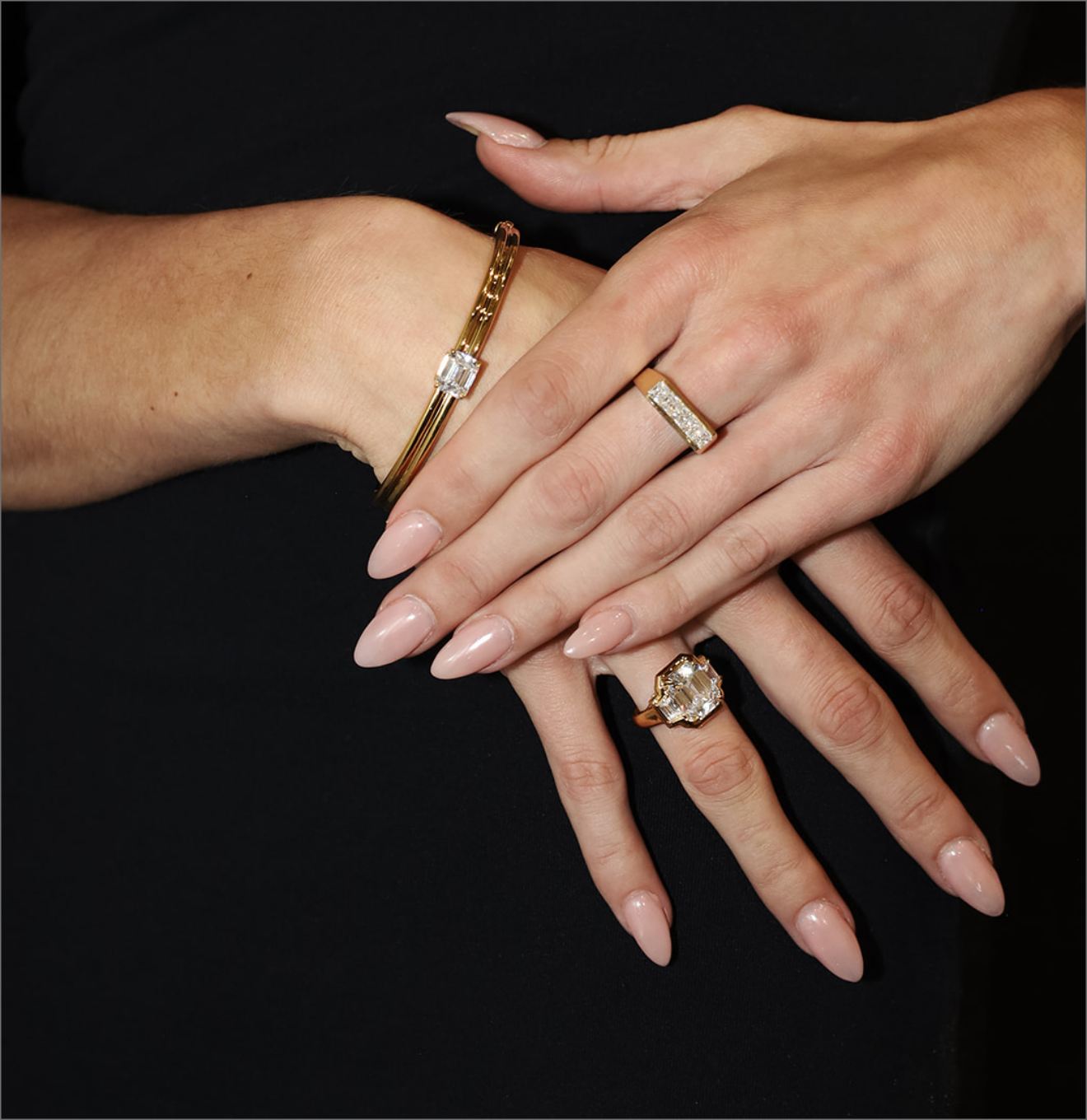 A close-up of crossed hands wearing gold jewelry, including a gold bracelet with a rectangular clear stone on one wrist, a gold ring with a clear rectangular stone on one finger, and a larger gold ring with a marquise-cut clear stone on the other finger, set against a solid black background.