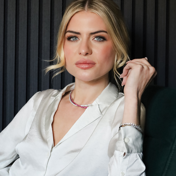 A blonde woman wearing a white satin button-up shirt, a pink and silver beaded necklace, and a silver bracelet, posing against a dark gray pleated background.