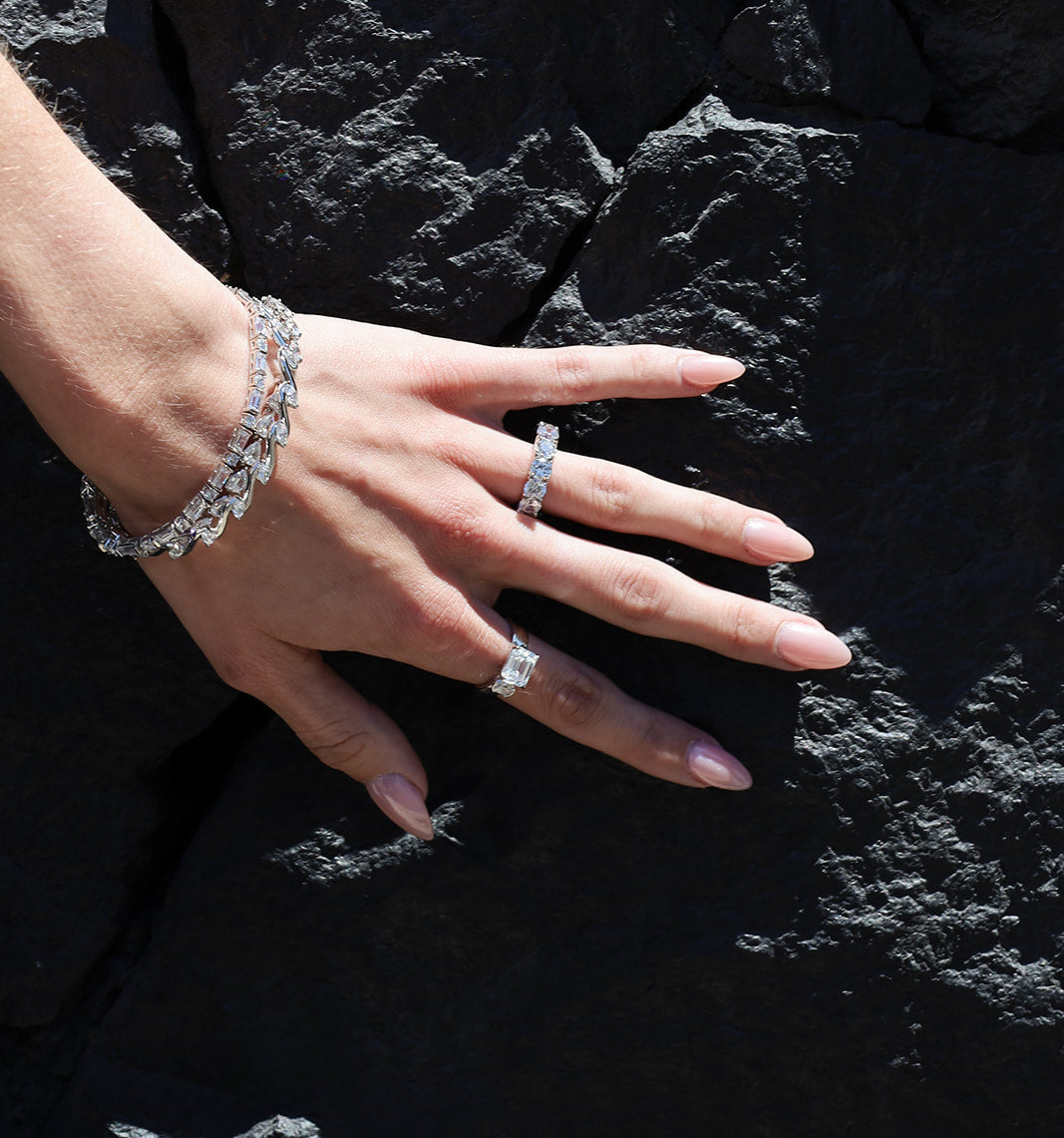 A close-up of a hand wearing a silver-toned diamond bracelet and matching diamond rings, posed against a dark textured rock surface.